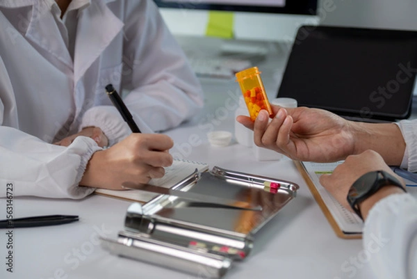 Fototapeta A doctor is writing in a notebook while a patient holds an orange pill bottle