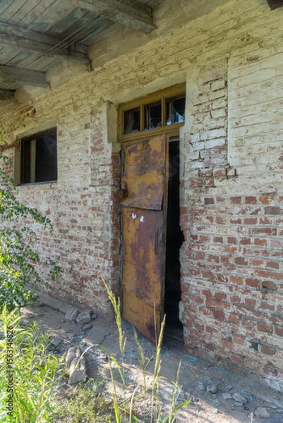 Fototapeta Abandoned brick building with rusted metal door slightly open, broken window panes, and crumbling facade, surrounded by overgrown vegetation under summer sunlight