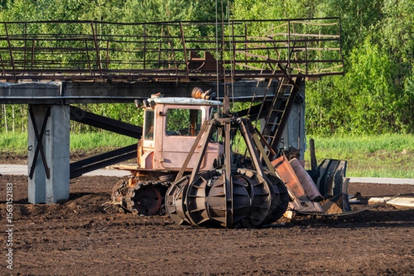 Fototapeta Old peat bog crawler excavator with large mechanical grabber bucket working under metal bridge structure, industrial machine on muddy brown field in rural Latvia landscape