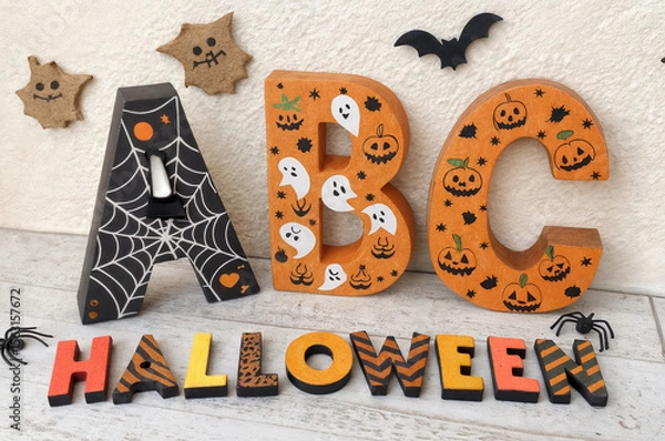 Fototapeta shows wooden letters and decorations with Halloween-themed designs, including spiders, bats, ghosts, and jack-o'-lanterns, arranged on a white wooden surface.