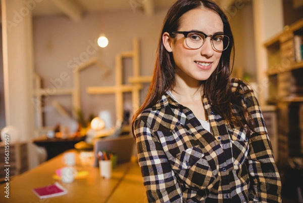 Obraz Woman standing behind wooden worktable in warm home office with mugs, notebooks, pens and laptop