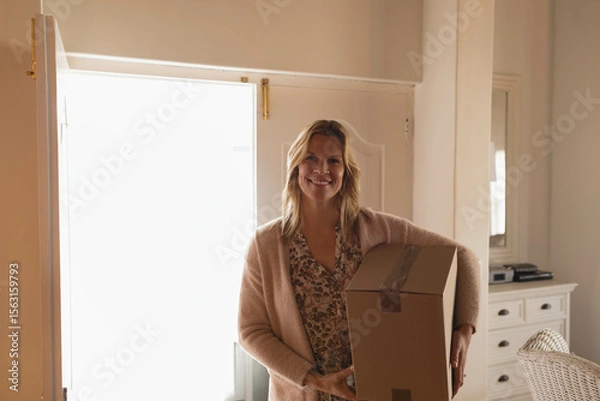 Obraz Woman standing near bright open doorway in entryway, holding moving box by dresser mirror, smiling