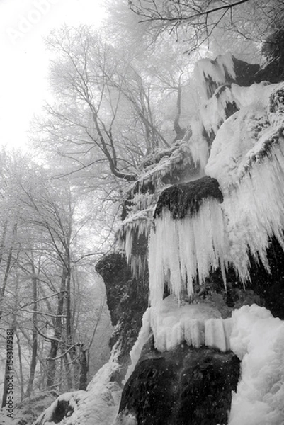Fototapeta “Uracher Wasserfall“ is a famous cascade and natural attraction near Stuttgart Germany. Many long icicles at frozen waterfall in winter season on a foggy day with snowfall. Black and white scenery.