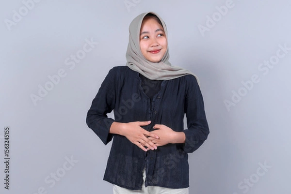 Fototapeta Smiling Asian Muslim woman in casual outfit and hijab posing against a plain background, looking aside with hands on stomach, expressing positive emotion.