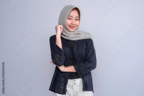 Fototapeta Portrait of a confident young Indonesian Muslim woman wearing a hijab and casual outfit, smiling with a hand on her chin, symbolizing intelligence, strength, and optimism.