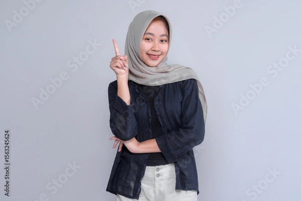 Fototapeta Cheerful young Indonesian Muslim woman in hijab smiling confidently while pointing upward with her index finger, symbolizing new ideas, awareness, solution, or inspiration.