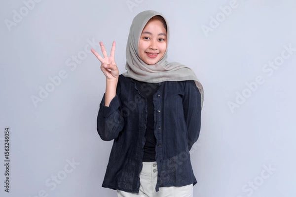 Fototapeta Portrait of a cheerful young Asian woman wearing a light grey hijab and black blouse, smiling confidently while showing three fingers gesture against a plain background.