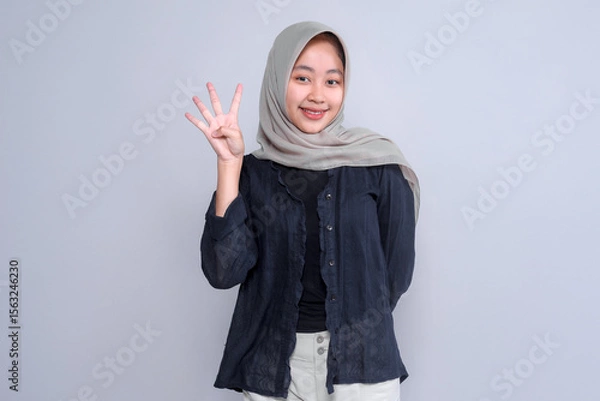 Fototapeta Portrait of a smiling young Indonesian woman in a hijab, showing four fingers with her right hand. She stands confidently against a plain grey background.