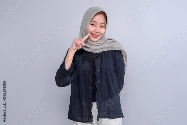 Fototapeta Portrait of a smiling young Indonesian woman wearing a grey hijab and black blouse, pointing to her cheek with one finger while standing confidently against a plain studio background.