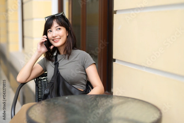 Obraz Happy young southeast asian woman blogger in 30s working outside room at coffee shop with old architecture searching internet on smartphone