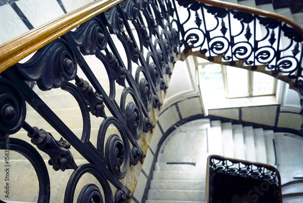 Fototapeta Beautiful curves and visual balance: architectural top-down view of spiral stairway with iron railing and marble steps. Life in an old European house: the beauty of the interior in a public space.