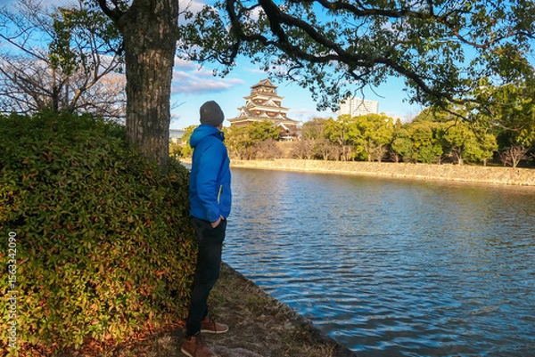 Fototapeta Male tourist in a blue jacket stands by the moat, admiring the view of iconic Hiroshima Castle. He enjoys a peaceful moment of contemplation while exploring the historic landmark in Hiroshima, Japan