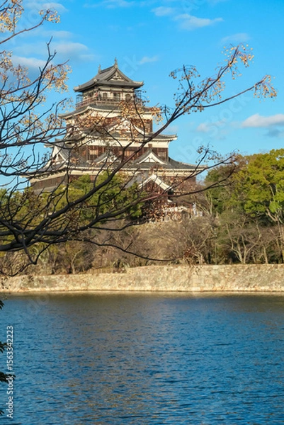 Fototapeta Historic Hiroshima Castle, known as the Carp Castle, rises majestically beyond its tranquil moat. This scenic view in Hiroshima, Japan, is framed by delicate tree branches against a clear blue sky