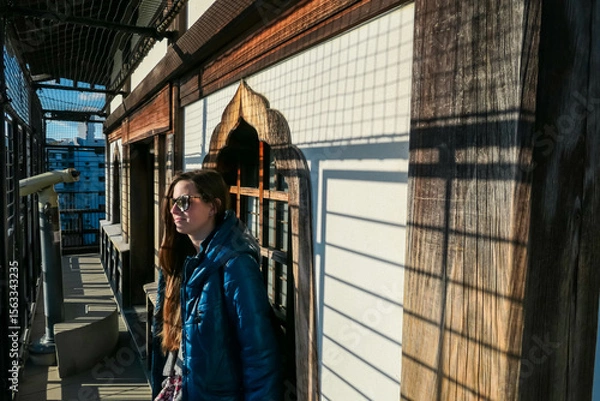Fototapeta Female tourist in sunglasses enjoys sun on the observation deck of Hiroshima Castle, Japan. Strong light casts a grid of shadows. Historic wooden architecture, creating unique, memorable travel moment