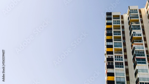 Fototapeta Tall residential apartments with geometric, colorful balconies under clear blue sky, copy space for text. Urban architecture image for real estate, housing, or city development context.