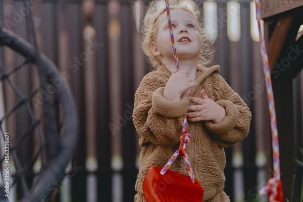 Fototapeta Beautiful 3-year-old girl playing on a neighborhood playground swinging, running on the grass, and enjoying fresh air. A joyful, natural moment full of real emotions and unaltered beauty