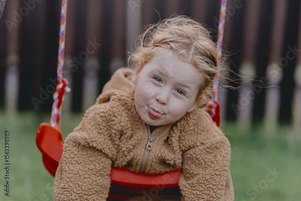 Fototapeta Beautiful 3-year-old girl playing on a neighborhood playground swinging, running on the grass, and enjoying fresh air. A joyful, natural moment full of real emotions and unaltered beauty