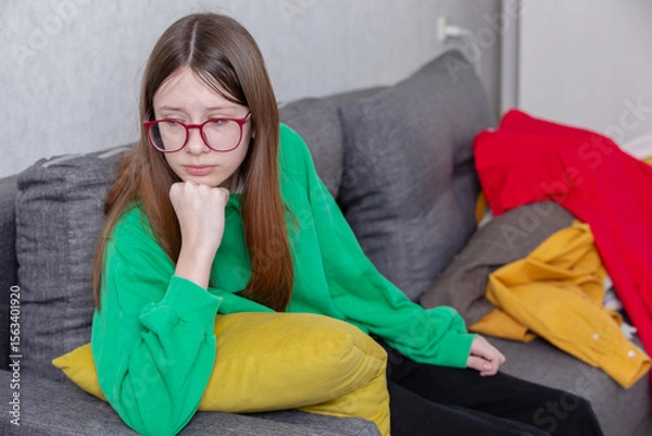 Obraz Teen girl in green sweatshirt sitting on gray couch with thoughtful expression and colorful clothes scattered around