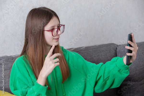 Obraz Teenager in green hoodie takes selfie while sitting on grey sofa in cozy indoor setting during afternoon