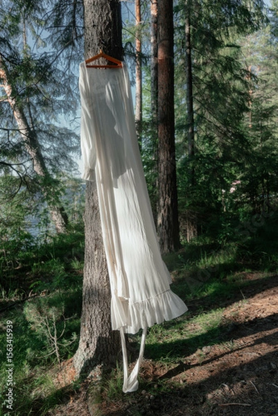 Fototapeta white dress is gracefully hung from a tree branch surrounded by lush greenery. Sunlight filters through the forest, creating a tranquil atmosphere in the afternoon