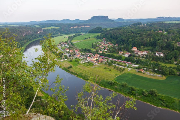 Obraz Scenic view over rural village of Rathen in Saxon Switzerland, Germany, with a red train passing through the Elbe valley, surrounded by forested hills, farmland, and sandstone cliffs in summer light.