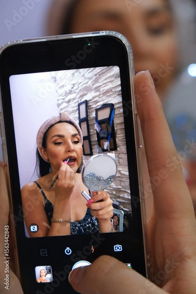 Fototapeta Taking a photo on a smartphone of a young beautiful woman applying lipstick. Everyday life. Concept of a woman's routine. beauty. self-care, blogging