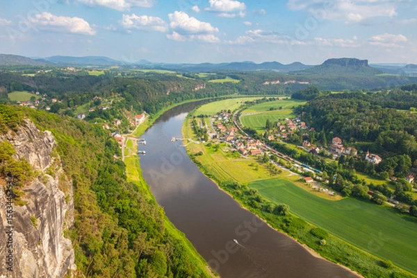 Obraz Scenic panoramic view from the Bastei cliffs over the Elbe River valley with sandstone rocks, lush forests, rural village rooftops, distant mountain ranges under a vibrant summer sky. Saxony, Germany