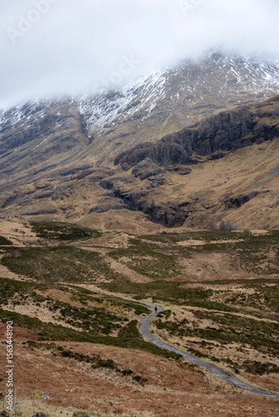 Fototapeta A solitary figure traverses the rugged, winding path through the majestic and mist-shrouded peaks of the Scottish Highlands, possibly in the iconic Glen Coe, where ancient landscapes meet dramatic ski