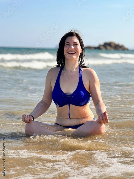 Fototapeta A smiling European woman in a blue bikini does yoga by the sea, sitting in a lotus position on the shore, with ocean waves in the background, vertical shot.