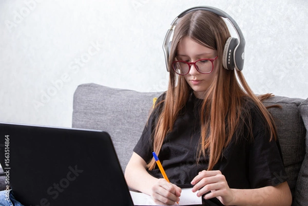Obraz Student engaged in online learning while taking notes at home with headphones on in a cozy environment