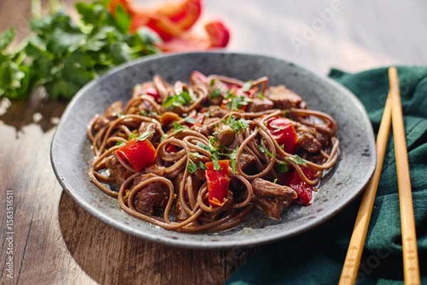 Fototapeta Wok with Soba noodles with turkey, mushrooms and sweet red bell pepper in plate on wooden table in the sun. Asian food, concept of street food.