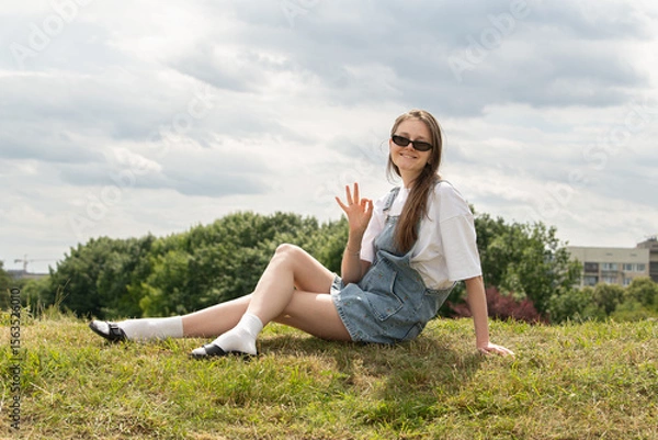 Fototapeta A young woman sits on a grassy hill, waving and smiling. She wears sunglasses and casual summer attire, enjoying a relaxed afternoon under a partly cloudy sky in an urban park