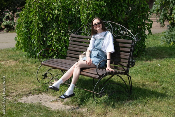 Fototapeta A woman relaxes on a metal bench in a vibrant garden, donning a casual outfit of a t-shirt and overalls. Sunlight filters through the trees, illuminating the scene