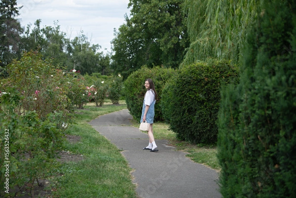 Fototapeta A young woman strolls along a winding path in a rose garden, surrounded by lush greenery and blooming flowers under a clear sky. The scene exudes a calm, serene atmosphere