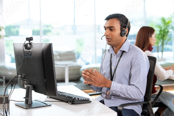 Fototapeta Professional tech support representative wearing headset provides customer assistance while working at modern office desk with computer, showcasing focused and helpful demeanor