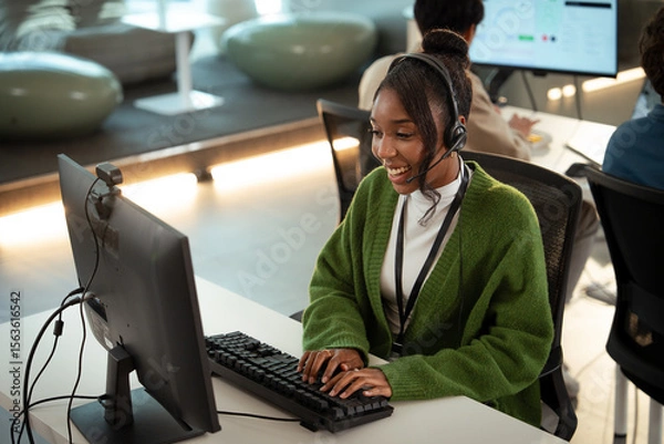 Fototapeta Cheerful customer support representative wearing headset and green sweater is working at computer in modern office environment, providing tech assistance