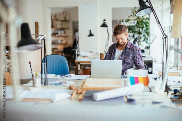 Fototapeta Smiling young architect working on laptop in creative office workspace