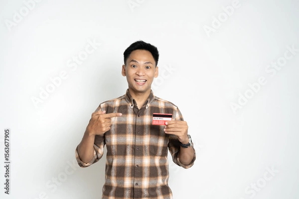 Fototapeta A cheerful young man proudly holding up and pointing to a credit card, symbolizing financial confidence, payment security, and personal identification in modern-day transactions.