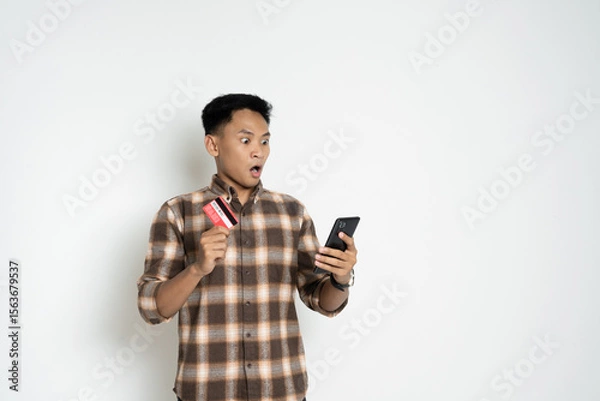 Fototapeta Young man in a brown plaid shirt looks shocked while holding a smartphone and credit card. Captures a concept of finance, online payment, digital transactions, and expressions of surprise or shock.