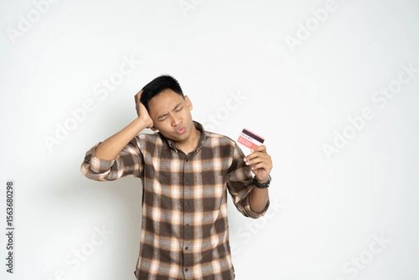 Fototapeta A young man expressing confusion while holding a credit card. Represents financial challenges, decision-making, or online transaction concerns.