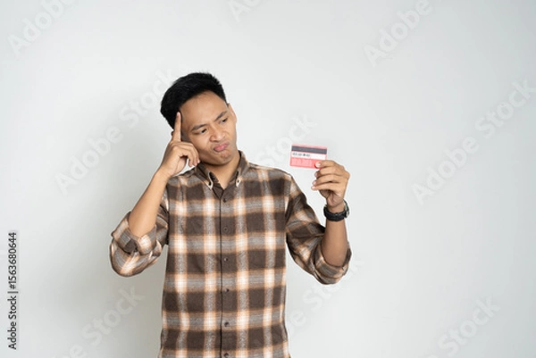 Fototapeta Young man in a brown plaid shirt looks shocked while holding a smartphone and credit card. Captures a concept of finance, online payment, digital transactions, and expressions of surprise or shock.