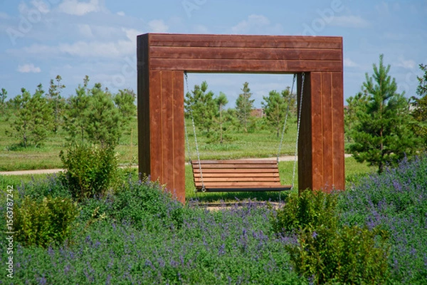 Obraz A wooden swing bench suspended within a minimalist timber frame, surrounded by purple flowering groundcover and young trees in a city park Photo