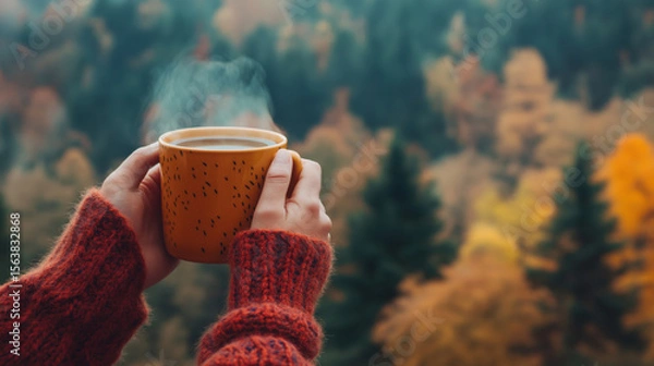 Fototapeta Hands in warm red sweater holding steaming cup against blurred autumn forest background with colorful trees. Cozy seasonal comfort and warmth.