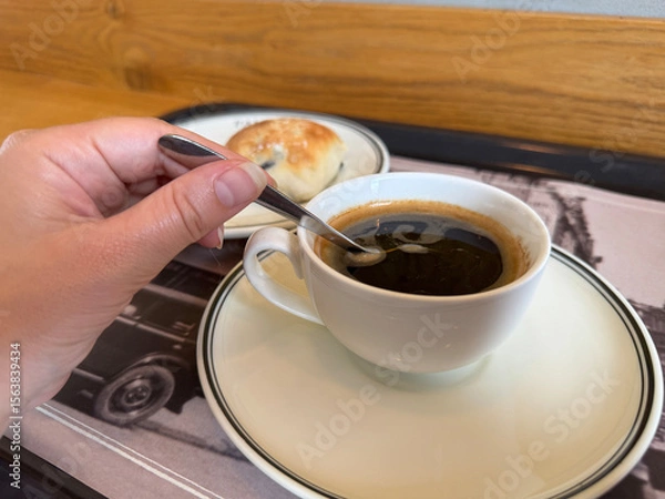 Fototapeta Cup of black coffee being stirred by hand with a spoon. Small bun on a side plate in cozy cafe setting