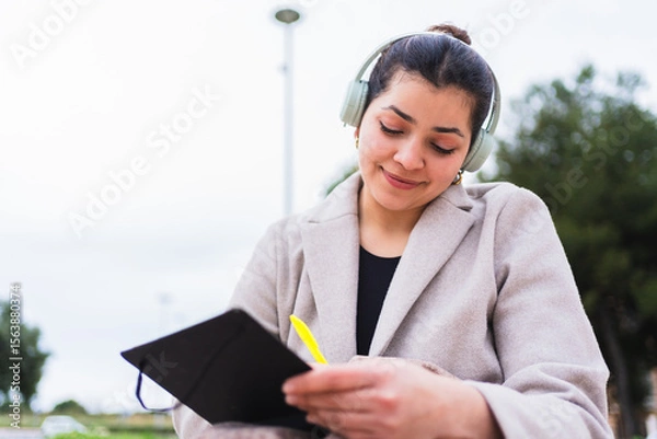 Fototapeta Young influencer writing notes on notebook while listening to music with headphones sitting in a park