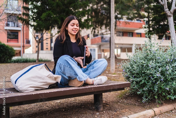 Fototapeta Young woman using digital tablet, creating online content while sitting on bench in city park