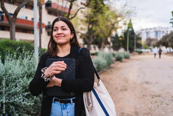Fototapeta Young influencer walking in a park, holding her tablet and smiling, enjoying a sunny day
