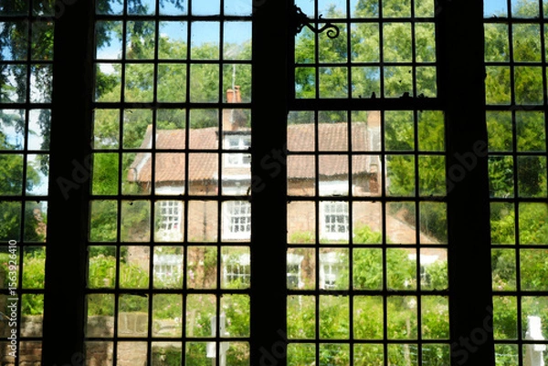 Obraz Oxton, Nottinghamshire / England - June 20 2025: Picturesque view through old church window to charming brick house with trees and blue sky