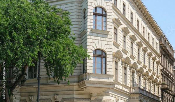 Fototapeta A corner view of a classic building with detailed stonework and large windows, surrounded by lush green trees.
