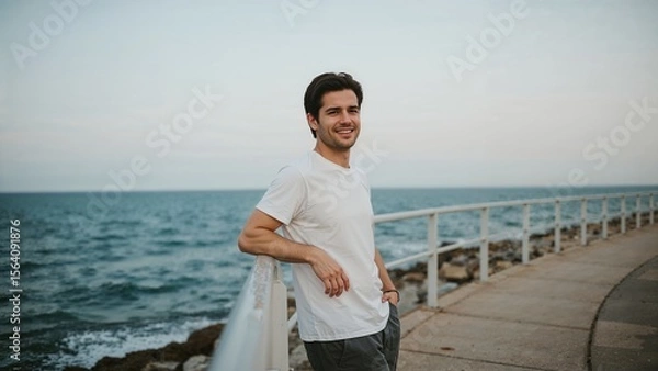 Fototapeta Smiling man enjoys ocean view from coastal pier, relaxing in casual wear on a bright day.